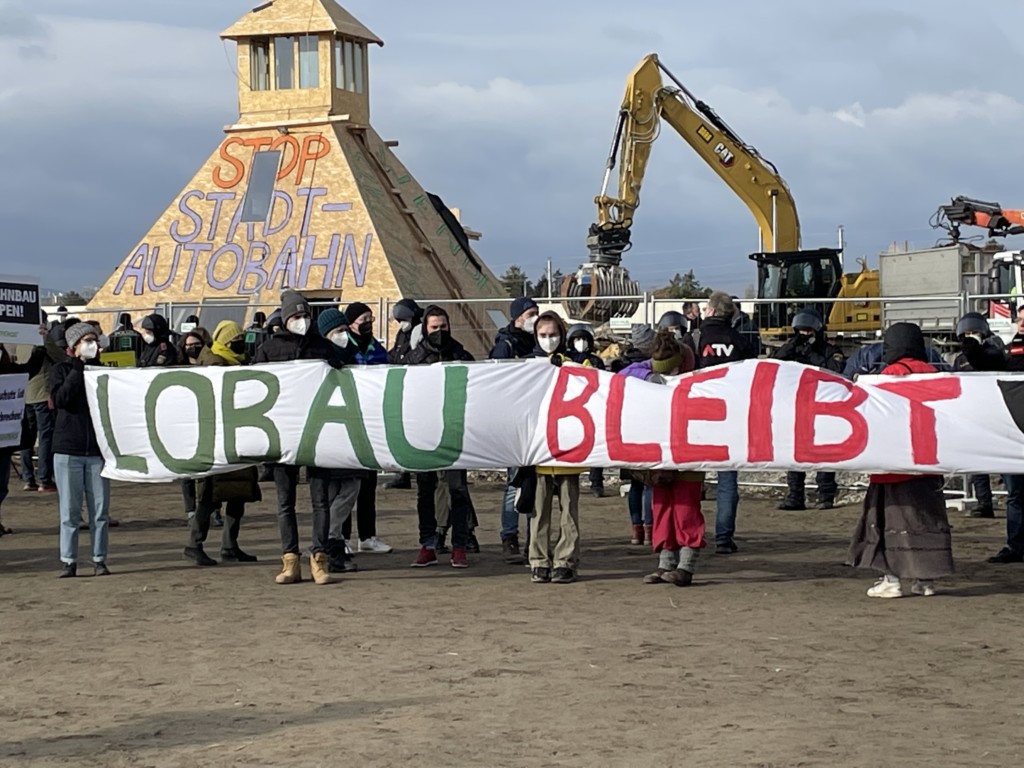 Räumungssituation im Wüstencamp, Menschen mit "Lobau bleibt"-Banner, im Hintergrund wird die Pyramide abgerissen